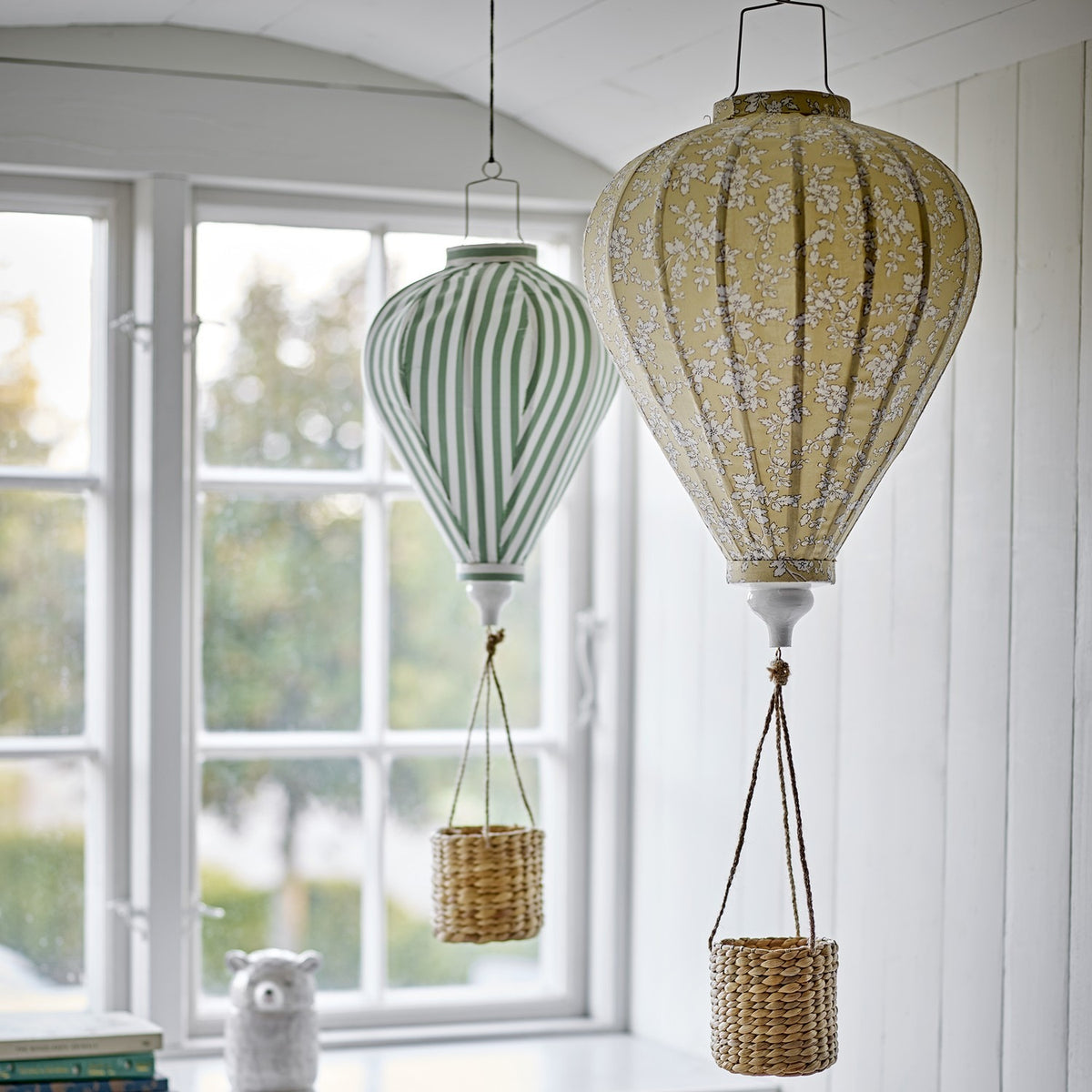 Pomp Floral Baby Mobile in yellow, a fabric hot air balloon-shaped hanging decor piece with a floral pattern and woven basket, shown beside a green striped balloon mobile near a window.