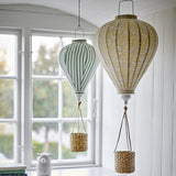Pomp Floral Baby Mobile in yellow, a fabric hot air balloon-shaped hanging decor piece with a floral pattern and woven basket, shown beside a green striped balloon mobile near a window.