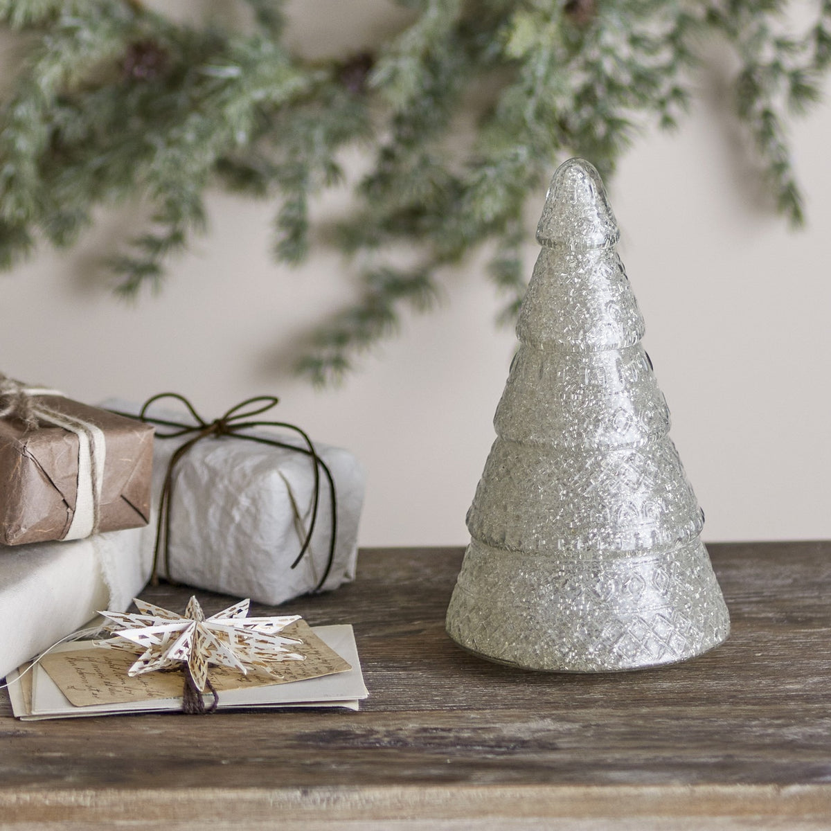 Emilius glass Christmas tree decoration in white, tiered fir shape with textured surface, displayed on a wooden surface beside wrapped gifts and greenery.