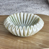 Harmonia Fluted Marble Bowl in white, 17.5cm, with deep even flutes and a wavy pleated rim, shown on a wooden surface against a grey textured backdrop.