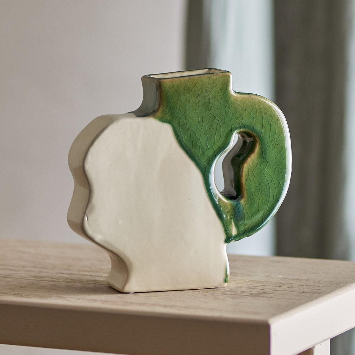 River Crackle Stoneware Vase 20cm in Green, a flat-sided ceramic vase with a wavy white profile and a green crackle-glazed handle, shown on a wooden table in a living room setting.