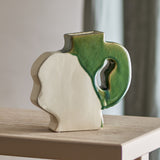 River Crackle Stoneware Vase 20cm in Green, a flat-sided ceramic vase with a wavy white profile and a green crackle-glazed handle, shown on a wooden table in a living room setting.