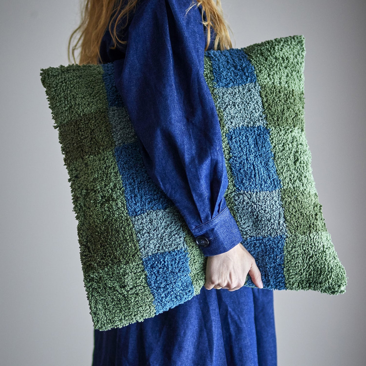 Pil Cotton Cushion in green, a square tufted cushion with a looped check pattern in green and blue, shown carried under the arm of a person in a blue outfit against a plain grey background.