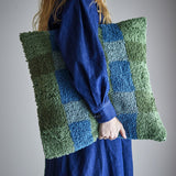 Pil Cotton Cushion in green, a square tufted cushion with a looped check pattern in green and blue, shown carried under the arm of a person in a blue outfit against a plain grey background.