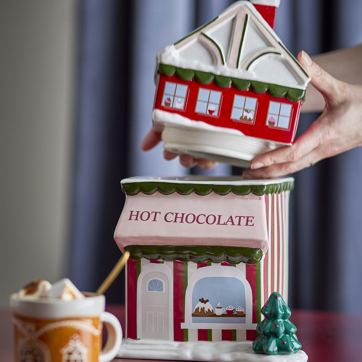 Gloria 2500ml ceramic Christmas storage jar shaped like a shop with HOT CHOCOLATE awning, lid lifted off, shown beside a mug of hot chocolate.