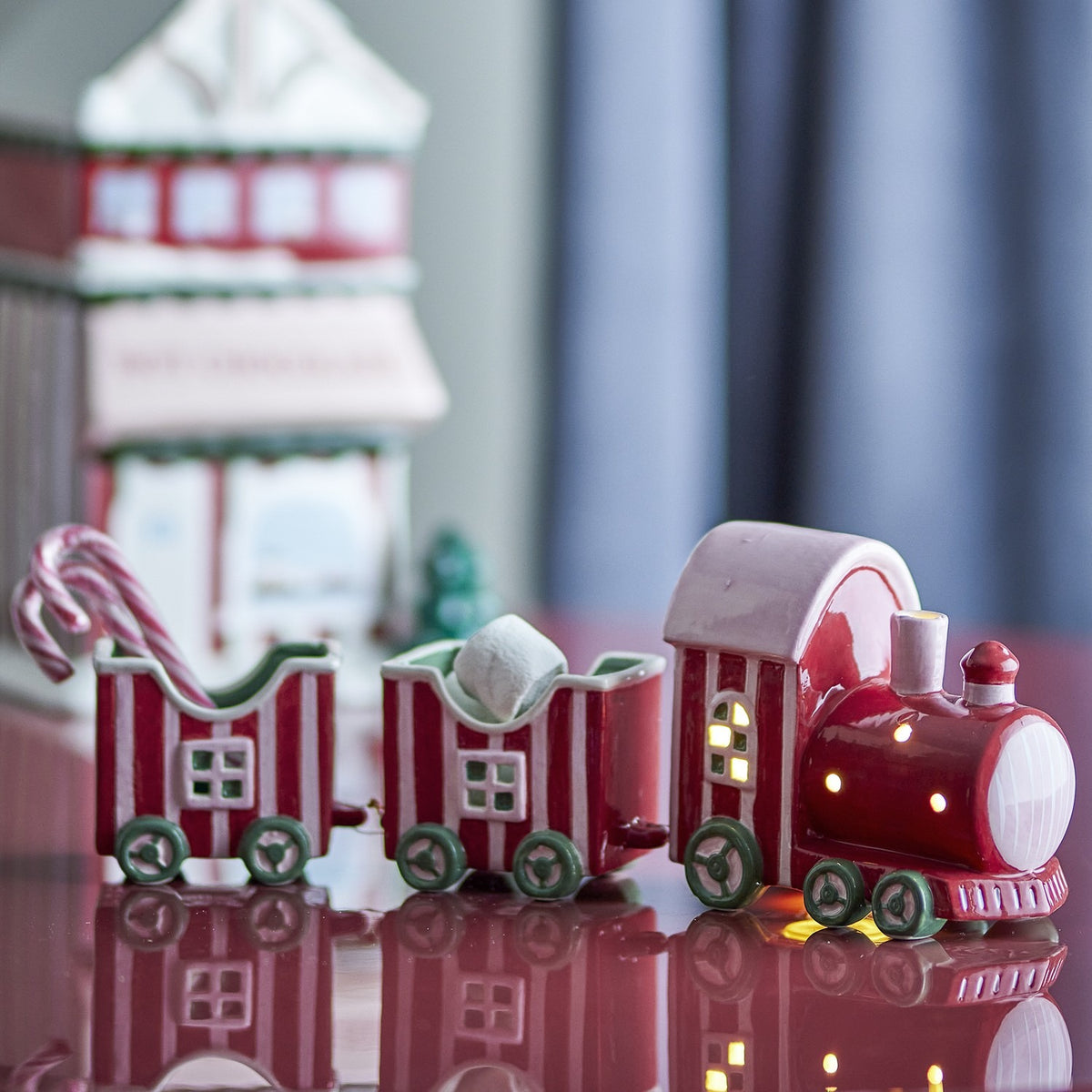 Gloria striped ceramic Christmas train decoration in red, with a chimney cab and two linked open wagons on green wheels, holding candy canes, shown on a reflective surface.