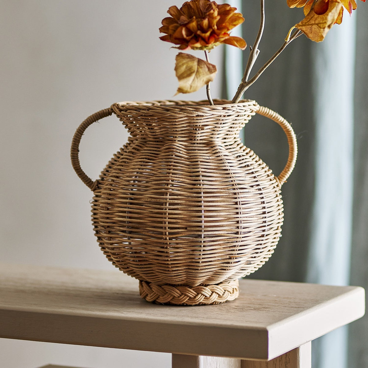 Uni Glass Vase 25.5cm in Brown, a round woven rattan urn-style vase with two curved handles, shown on a wooden table holding dried autumn florals.