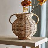 Uni Glass Vase 25.5cm in Brown, a round woven rattan urn-style vase with two curved handles, shown on a wooden table holding dried autumn florals.