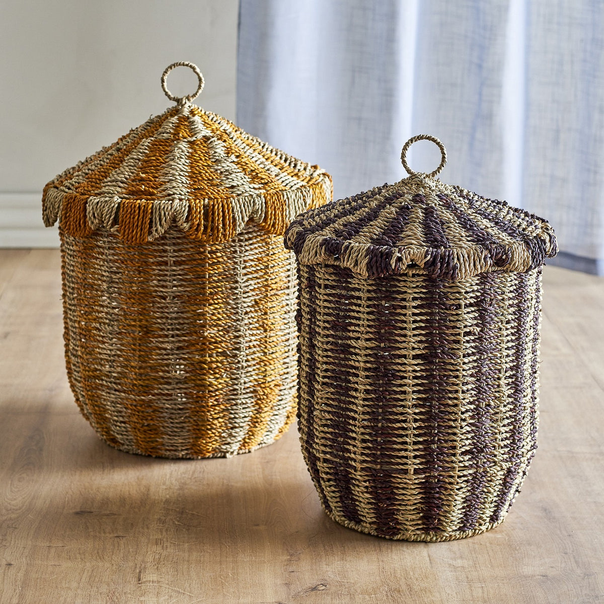 Cikkeline Striped Seagrass Baskets with Lids, set of two, tapered tent-shaped lids with scalloped trim and woven loop handles, in orange and purple striped seagrass, shown on a wooden floor.