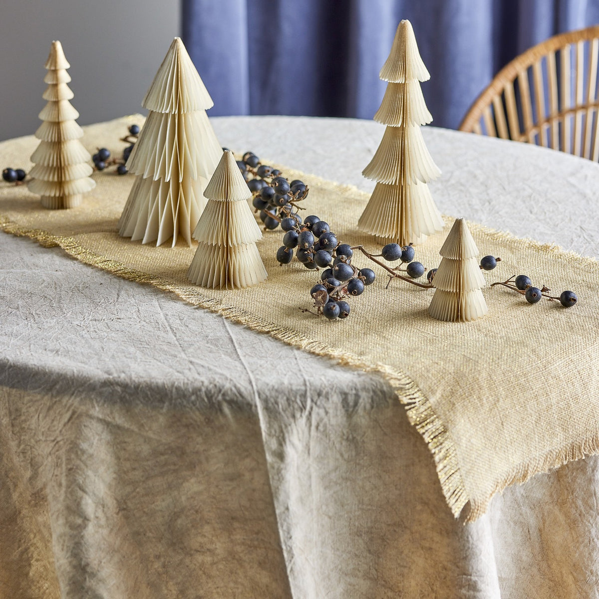 Milan Paper Christmas Decorations Set of 5 in cream, five tiered folded-paper trees of varying heights styled on a table runner with dark berries, a pale tablecloth and blue curtain behind.