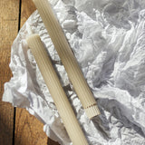 Ponza LED candles, Pack of 2 in Beige, tall ribbed paraffin-wax columns with flame-shaped tips, laid on crumpled white paper over a wood surface.