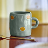 Agnes Stoneware Cup 230ml in Green with a glossy, uneven surface and hand-painted lemon motifs, shown with its handle on a wooden table beside a gold spoon.