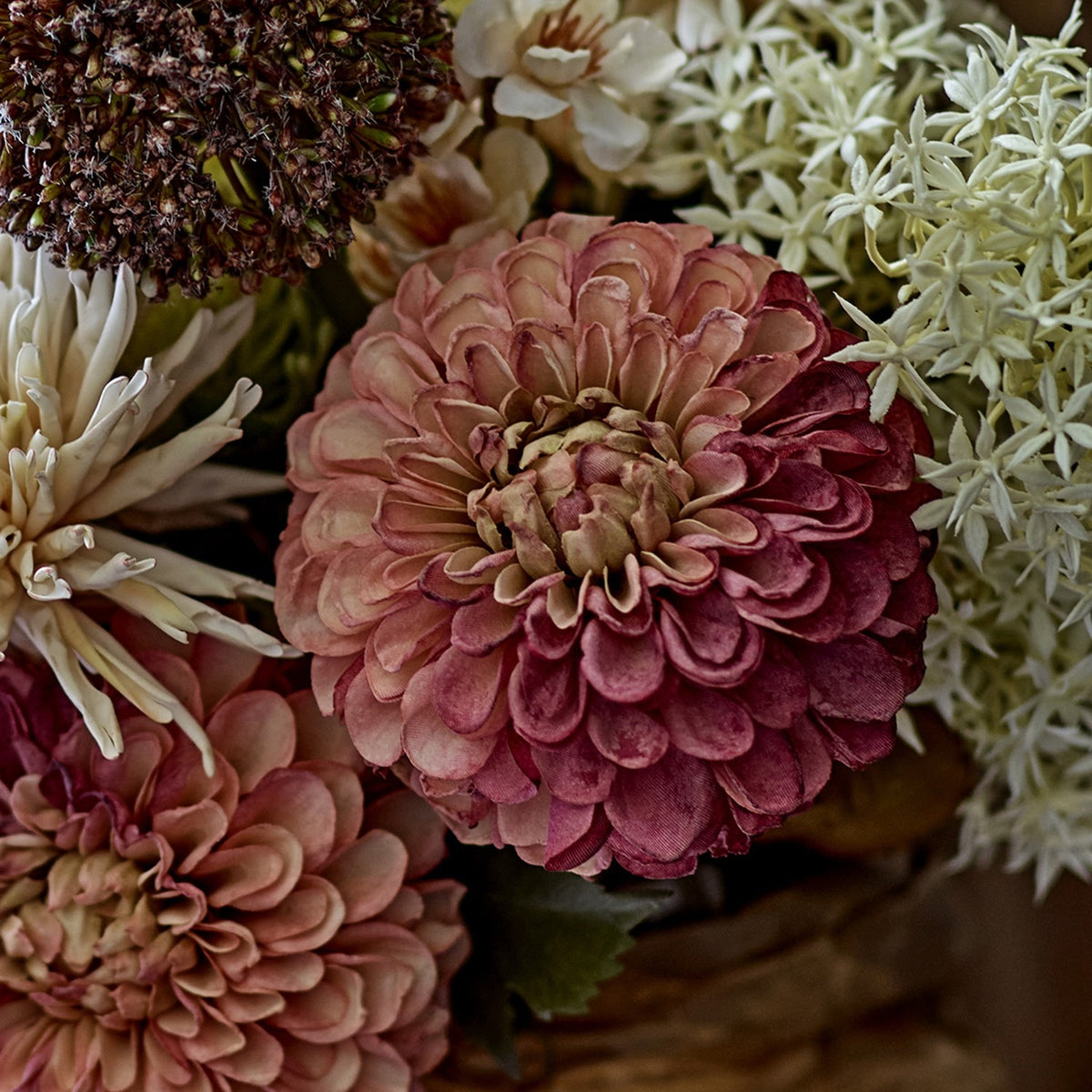 Artificial chrysanthemum stem with a full, rounded pink flower head in layered cupped petals, shown close-up among other dried-style blooms in warm pink and cream tones.