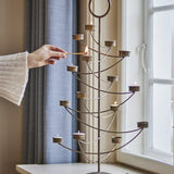 Alani Iron Christmas Tealight Holder in gold, tree-shaped with tiered curved arms and tealight cups, shown lit beside a window as a hand holds a match.