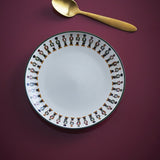 Jolly Stoneware Christmas Plate in cream with a ring of colourful nutcracker figures around the rim and a green edge line, shown beside a gold spoon on a dark red surface.