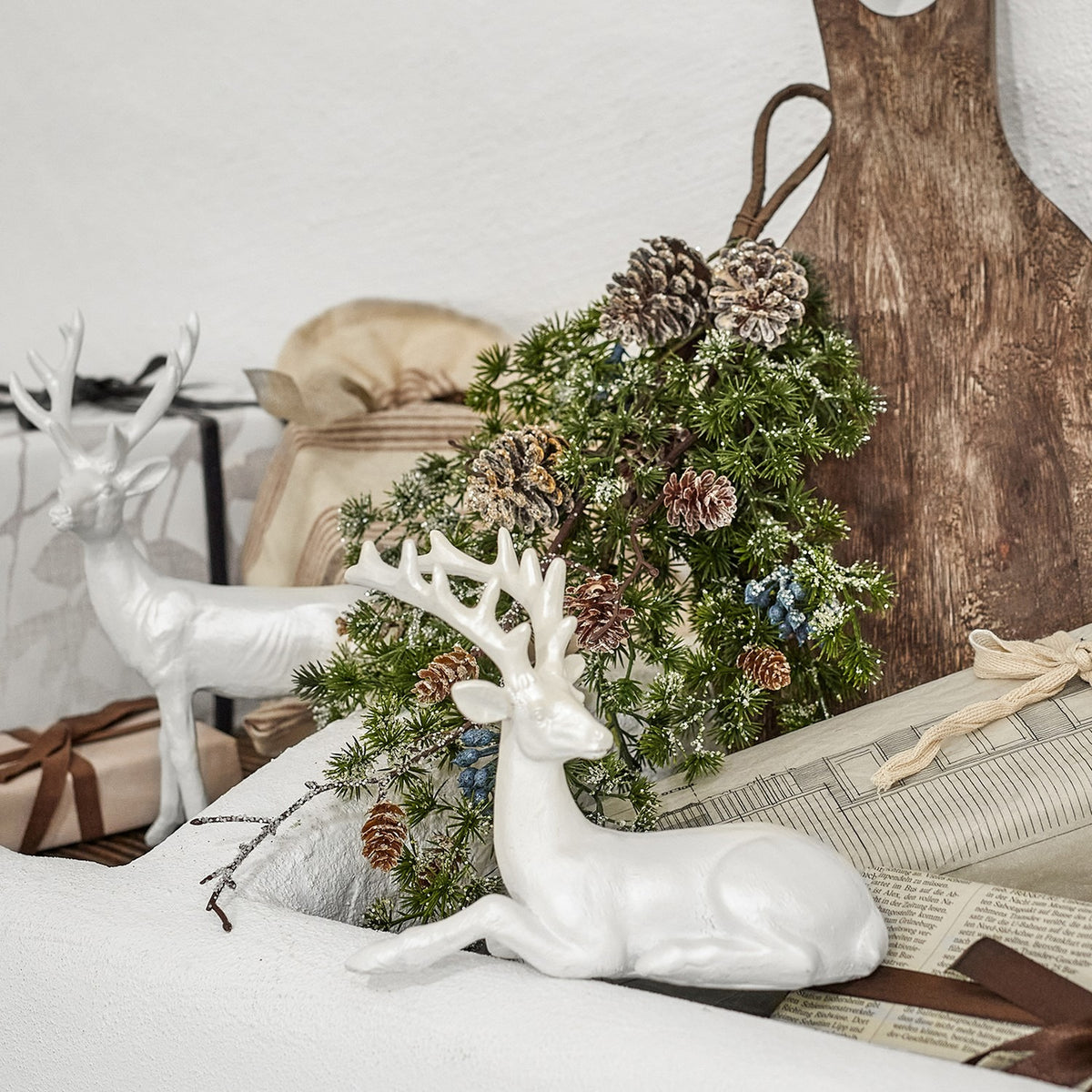 Cedar Christmas Decoration in green with frosted pinecones and berry clusters, displayed beside white ceramic reindeer figurines on a mantel.