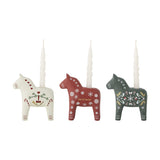Set of 3 resin horse-shaped Christmas candle holders in white, red and green, each hand-painted with hearts, snowflakes or folk motifs, shown with taper candles on a plain background.