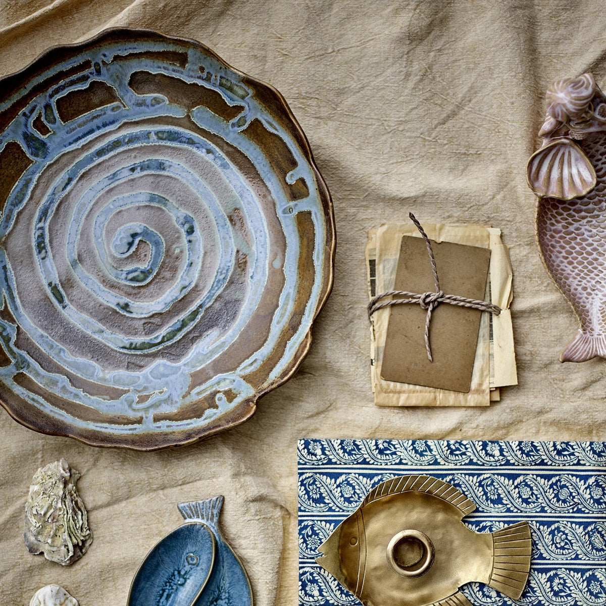 Marjorie Round Stoneware Tray in brown with a spiral glaze pattern and scalloped rim, shown among ceramic fish dishes, a brass fish dish, and paper ephemera on a linen cloth.