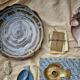 Marjorie Round Stoneware Tray in brown with a spiral glaze pattern and scalloped rim, shown among ceramic fish dishes, a brass fish dish, and paper ephemera on a linen cloth.