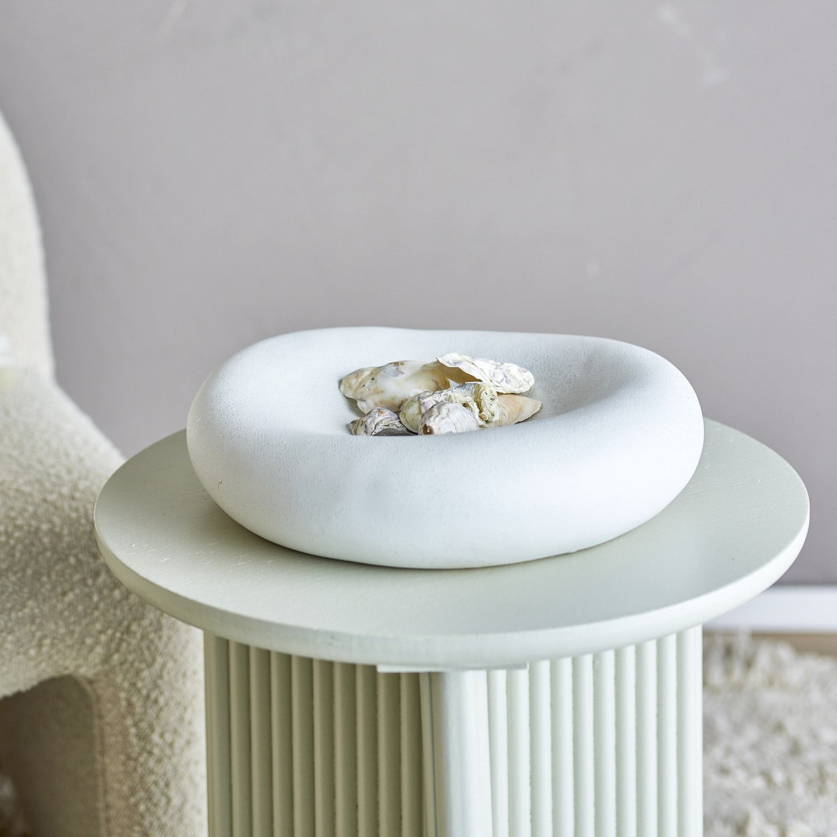 Audra Pebble Stoneware Bowl 30cm in White, a round pebble-shaped dish with a speckled finish holding seashells, displayed on a ridged pedestal side table.