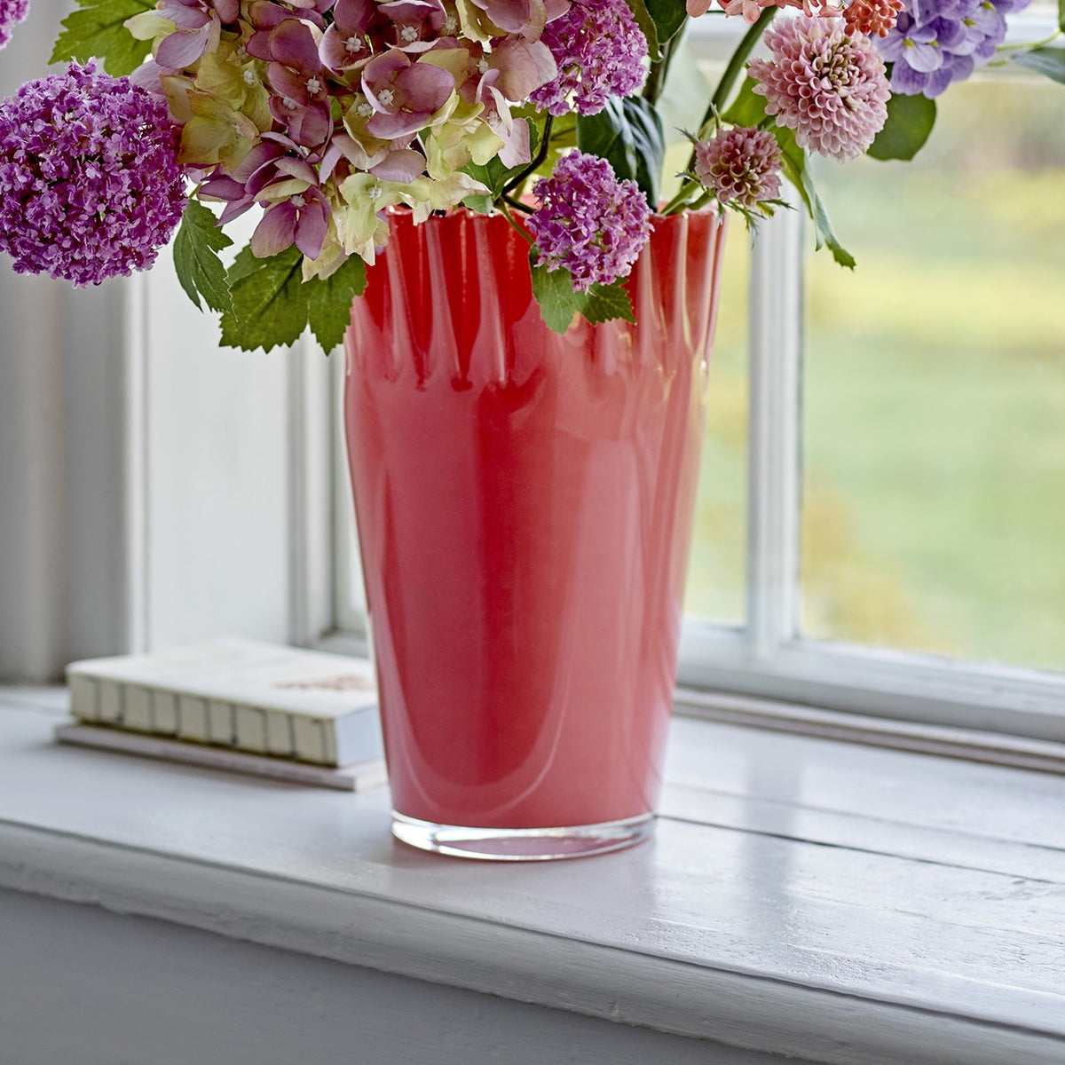 Janine Fluted Glass Vase 30cm in pink, with wavy frilled rim and tapered body, holding a mixed floral bouquet on a white windowsill.