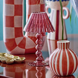 Cisco Glass Tealight Holder in pink, shaped like a table lamp with a fluted, wavy-rimmed shade atop stacked glass beads, displayed on a table among striped vases and a checkered jug.