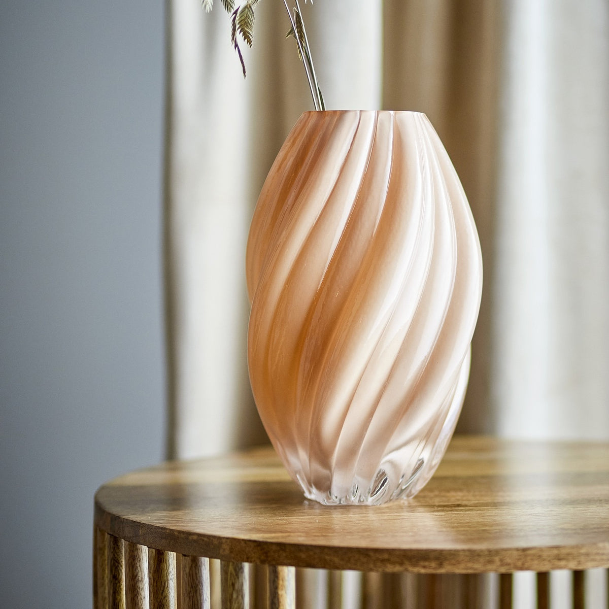 Kanji Glass Vase 30cm in Beige, a spiral-fluted glass vase with a swelling body and narrow rim, shown with foliage on a wooden side table against a curtained backdrop.