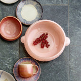 Lalie Ribbed Glass Bowl in pink, 20x12.5cm, with handled lugs and ribbed exterior, shown on a tiled surface alongside patterned plates, a mug, and a bowl filled with redcurrants.
