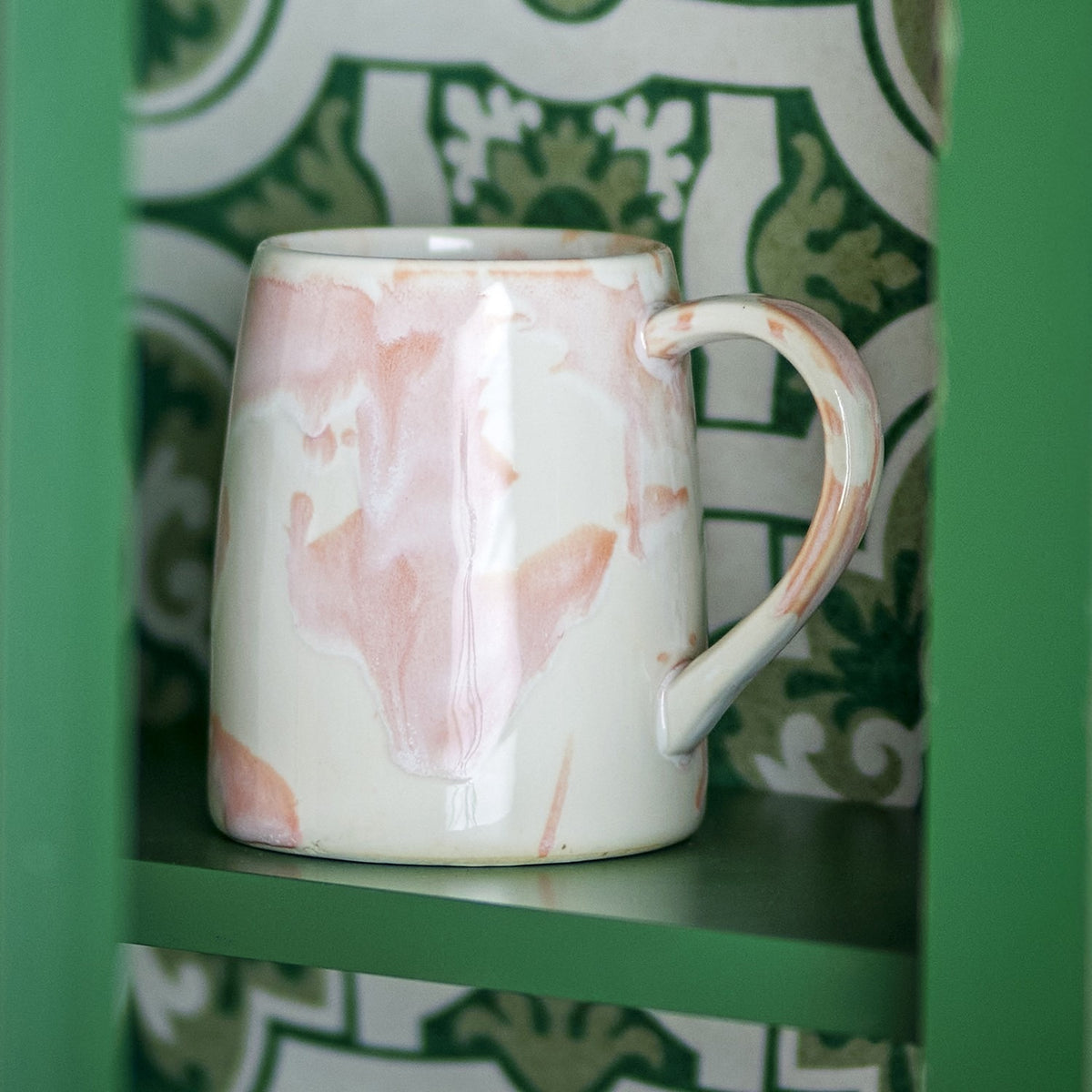 Winnie Marbled Stoneware Mug in pink and white reactive glaze with open loop handle, shown on a green shelf against a green and white patterned backdrop.