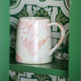 Winnie Marbled Stoneware Mug in pink and white reactive glaze with open loop handle, shown on a green shelf against a green and white patterned backdrop.