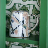 Winnie Splattered Stoneware Mug in multicoloured reactive glaze with drips and speckles over the body and curved handle, shown on a green shelf against patterned green and white tiles.