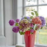 Edge Artificial Bouquet in Purple displayed in a red vase on a windowsill, featuring hydrangea-style clusters, pompom heads, and trailing leaves in shades of purple, pink, and green.