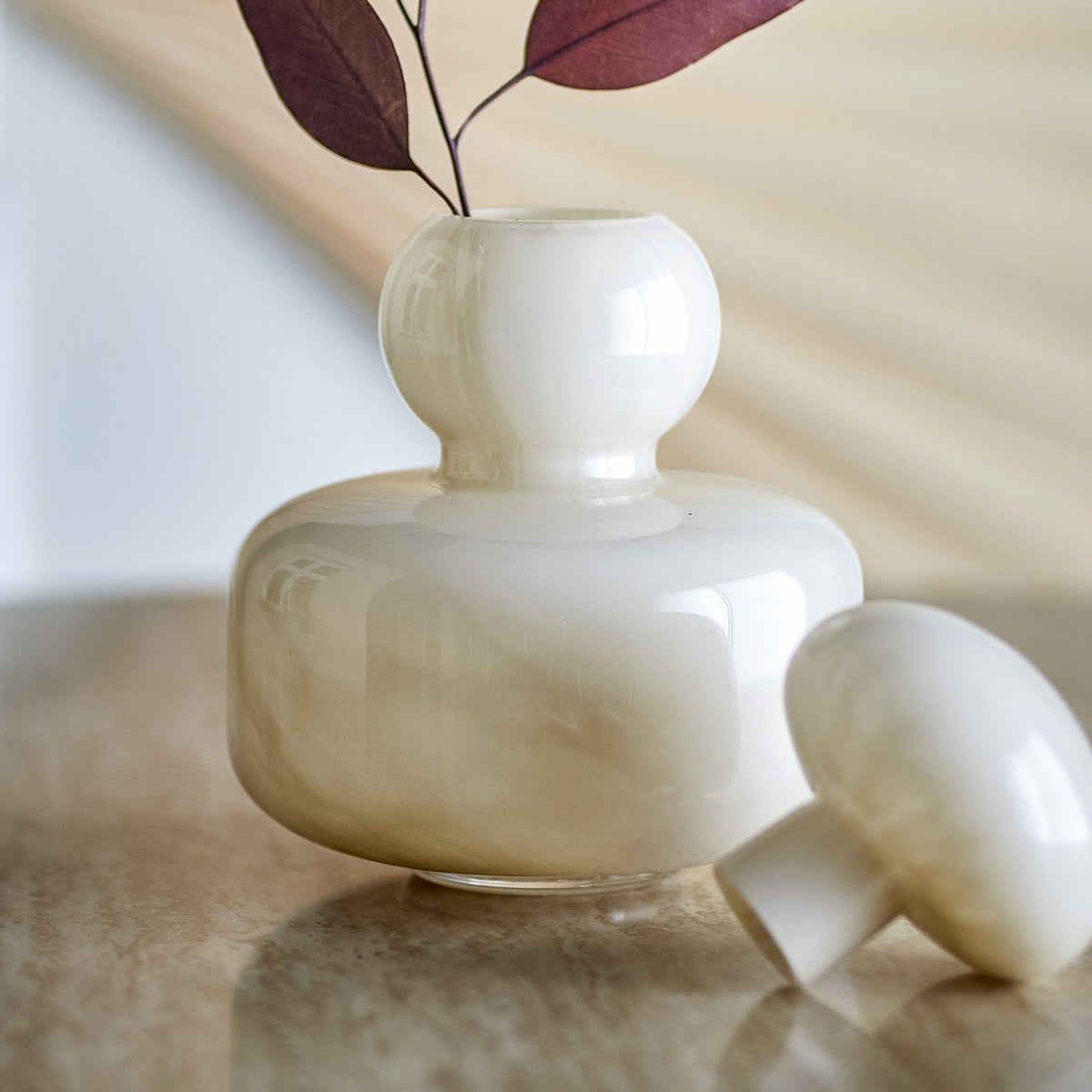 Cynthia Glass Vase 21cm in Beige, mouth-blown glossy glass with rounded body and ball-shaped neck, domed mushroom-style stopper resting beside it on a marble surface.