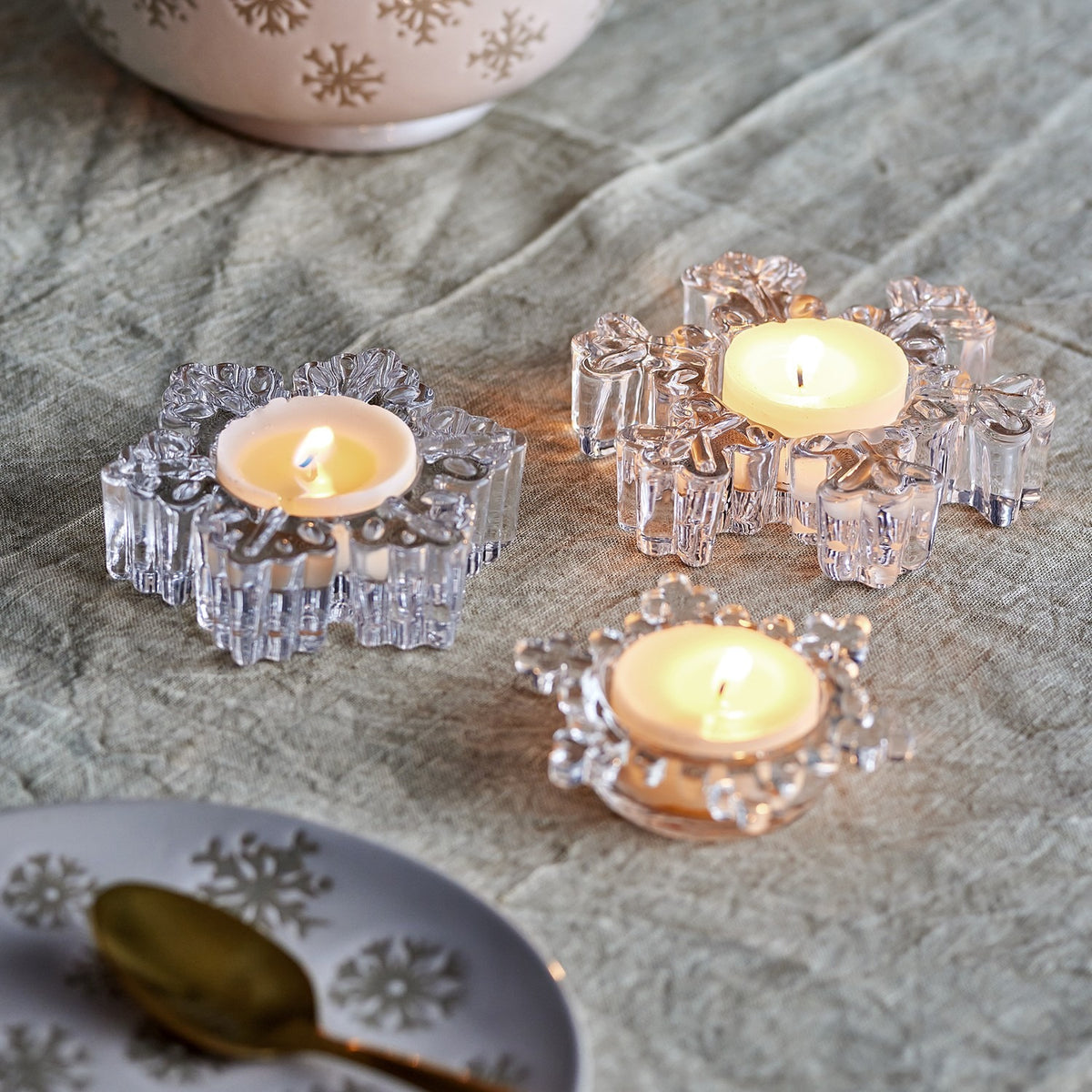 Dania Snowflake Glass Christmas Tealight Holders, Set of 3 in Clear, each uniquely shaped with lit tealights, styled on a grey fabric tabletop with plates and a bowl.