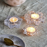 Dania Snowflake Glass Christmas Tealight Holders, Set of 3 in Clear, each uniquely shaped with lit tealights, styled on a grey fabric tabletop with plates and a bowl.