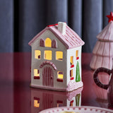 Shirly House stoneware Christmas tealight holder in white, with a pink pitched roof, chimney, arched door and cut-out windows glowing from an inner light.