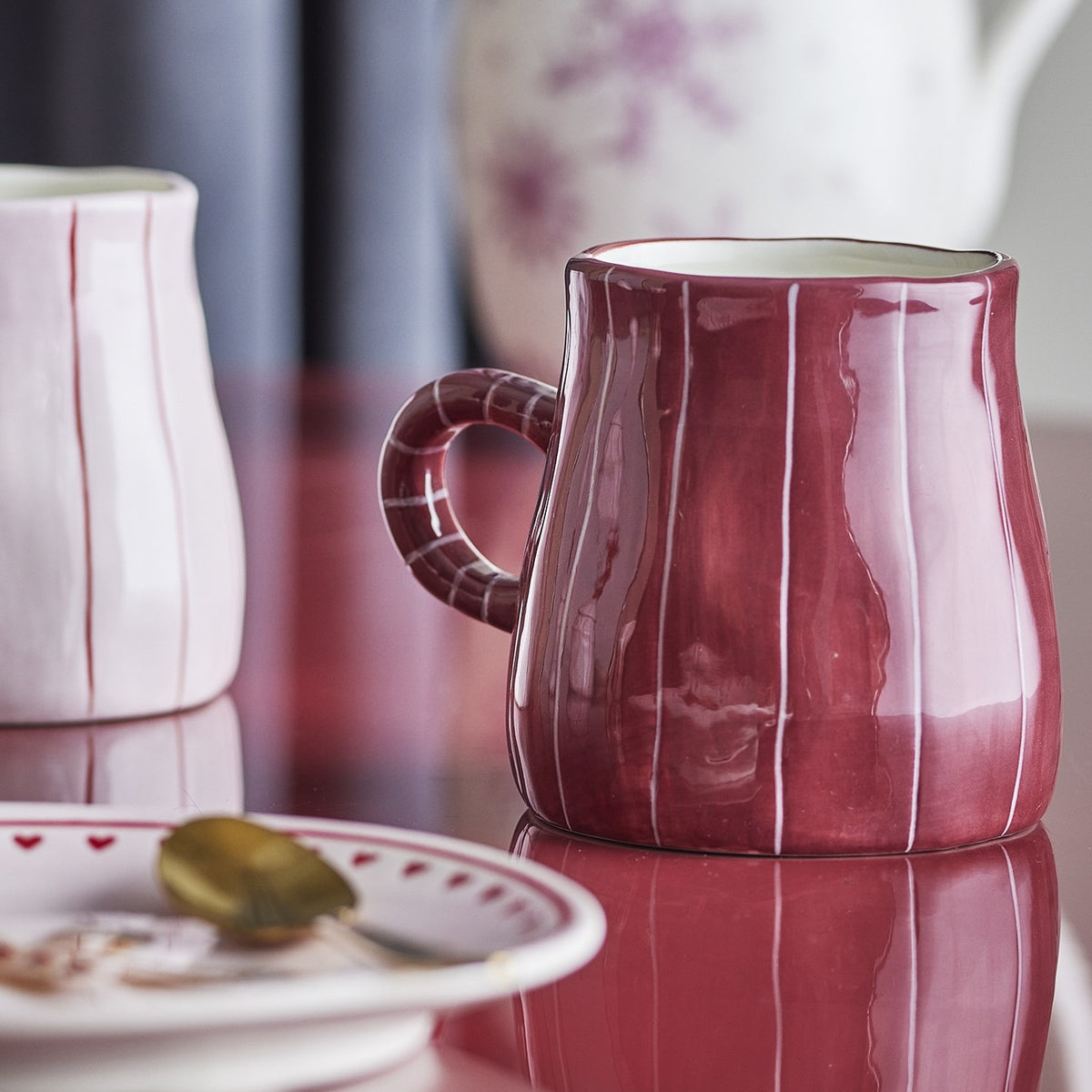 Shirly Striped Stoneware Christmas Mug 260ml in deep red with fine white stripes and a ring-shaped handle, shown on a glossy surface with its pale pink striped pair blurred behind.