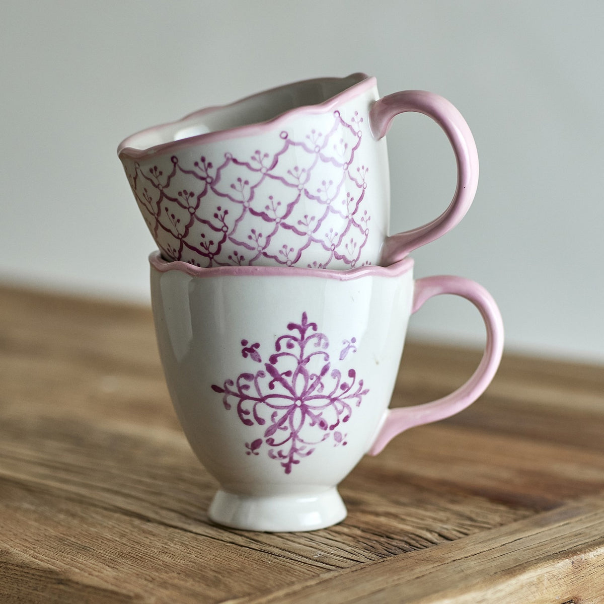 Two stacked stoneware mugs from the Wonderland Christmas set in white with pink scalloped rims and handles, one with a trellis pattern, one with a snowflake motif, on a wooden surface.