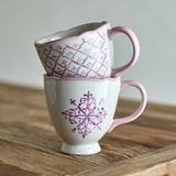 Two stacked stoneware mugs from the Wonderland Christmas set in white with pink scalloped rims and handles, one with a trellis pattern, one with a snowflake motif, on a wooden surface.