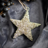 Sarika Star Iron Christmas Tree Decoration in gold with a textured, crumbly finish and jute string hanging loop, shown on a dark grey fabric backdrop with black berries.