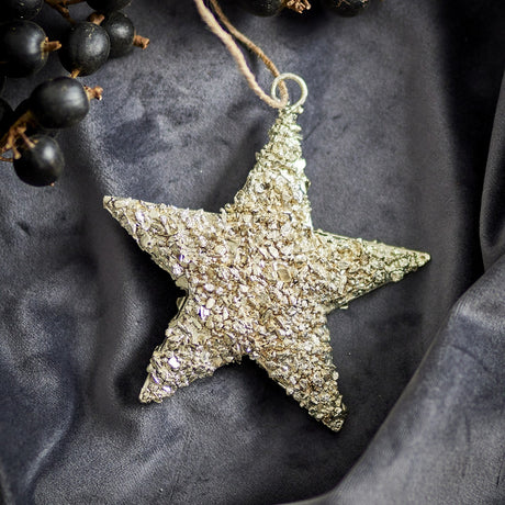 Sarika Star Iron Christmas Tree Decoration in gold with a textured, crumbly finish and jute string hanging loop, shown on a dark grey fabric backdrop with black berries.