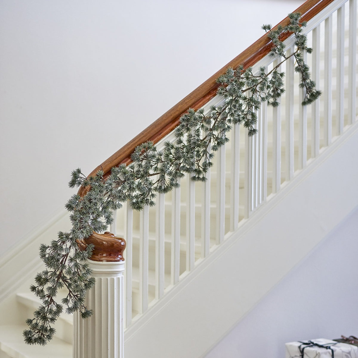 Artificial cedar Christmas garland in frosted green draped along a white staircase handrail with wooden rail cap and turned newel post.