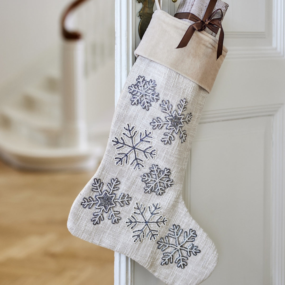 Beige woven Ahsan Christmas stocking with a folded cuff and beaded snowflake embroidery, hung on a white door in a hallway setting.