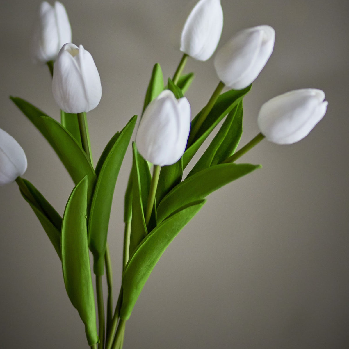 Artificial bouquet of white tulips with closed petals on tall stems and pointed green leaves, set against a plain grey background.