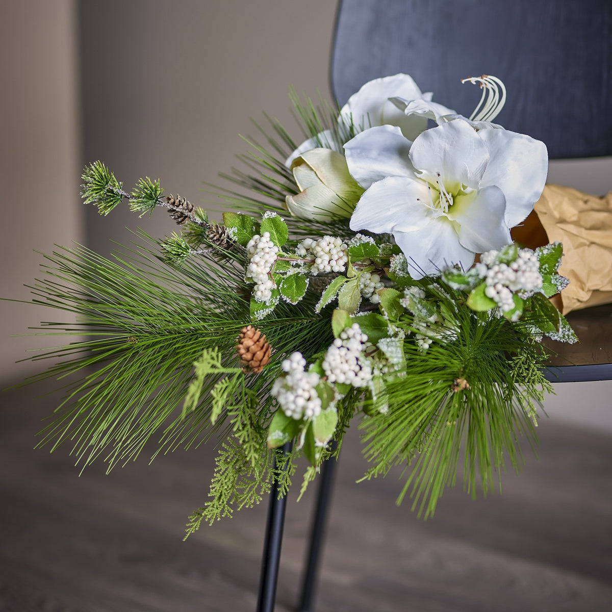 Kristin Artificial Christmas Bouquet in White, with large trumpet-shaped blooms, pine needles, frosted leaves and white berry clusters, displayed on a chair indoors.