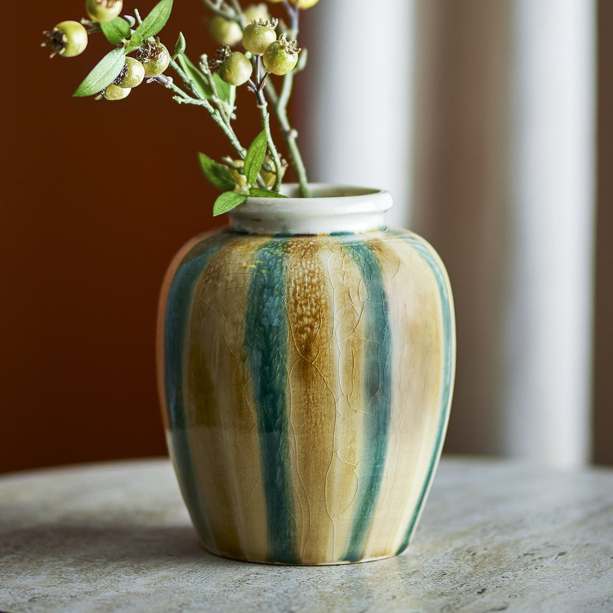 Rahima Crackle Stoneware Vase 20cm with hand-painted teal and ochre stripes and a crackle glaze, round body and white rim, holding a leafy berry sprig on a marble surface.