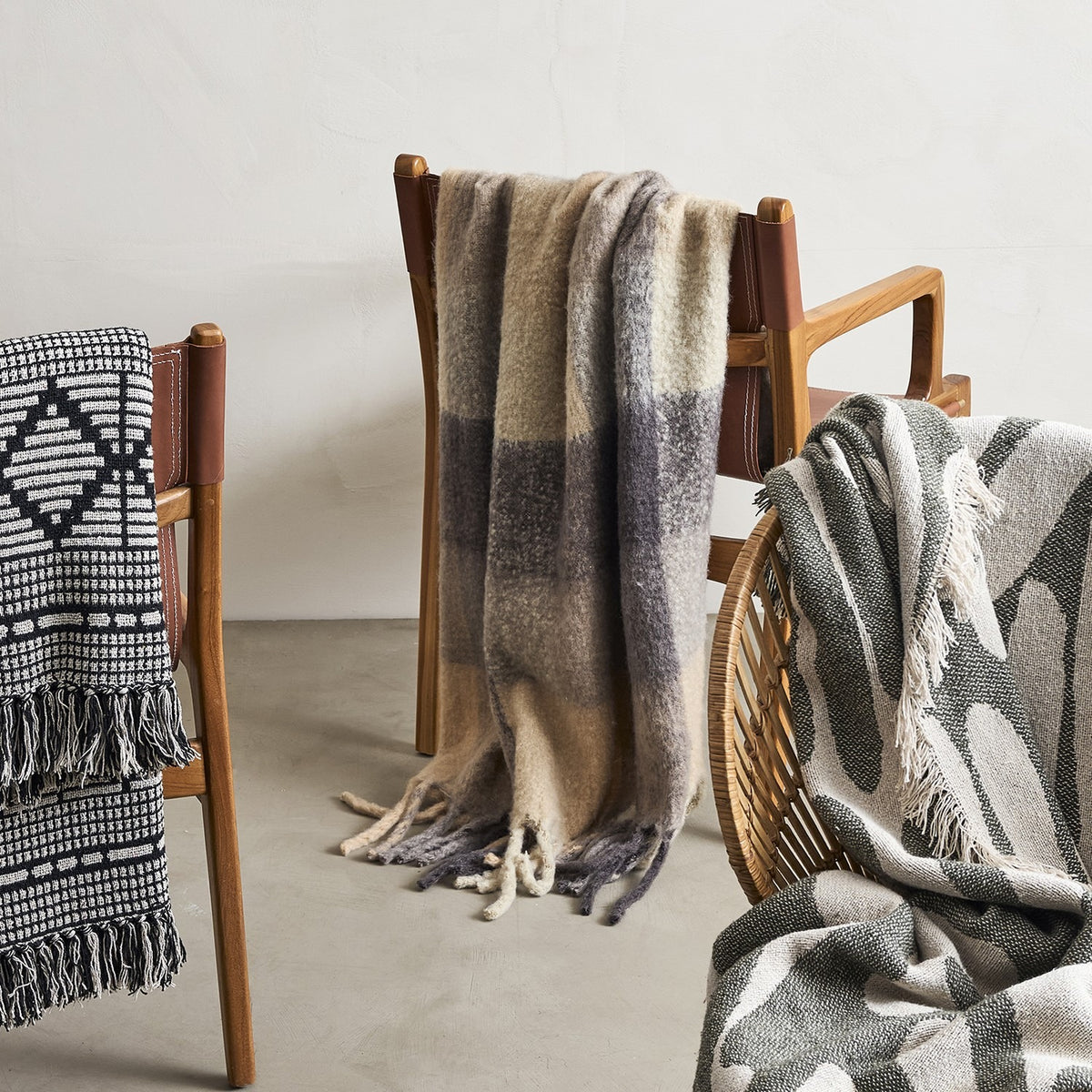 Hamisha Grey Throw in a multicoloured block check with fuzzy texture and long fringe, draped over a wooden chair alongside other patterned throws.
