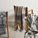 Hamisha Grey Throw in a multicoloured block check with fuzzy texture and long fringe, draped over a wooden chair alongside other patterned throws.