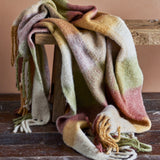 Hamisha Throw in Multi, a woven blanket-check throw with twisted fringe in pink, gold and green, draped over a wooden stool against a terracotta wall.