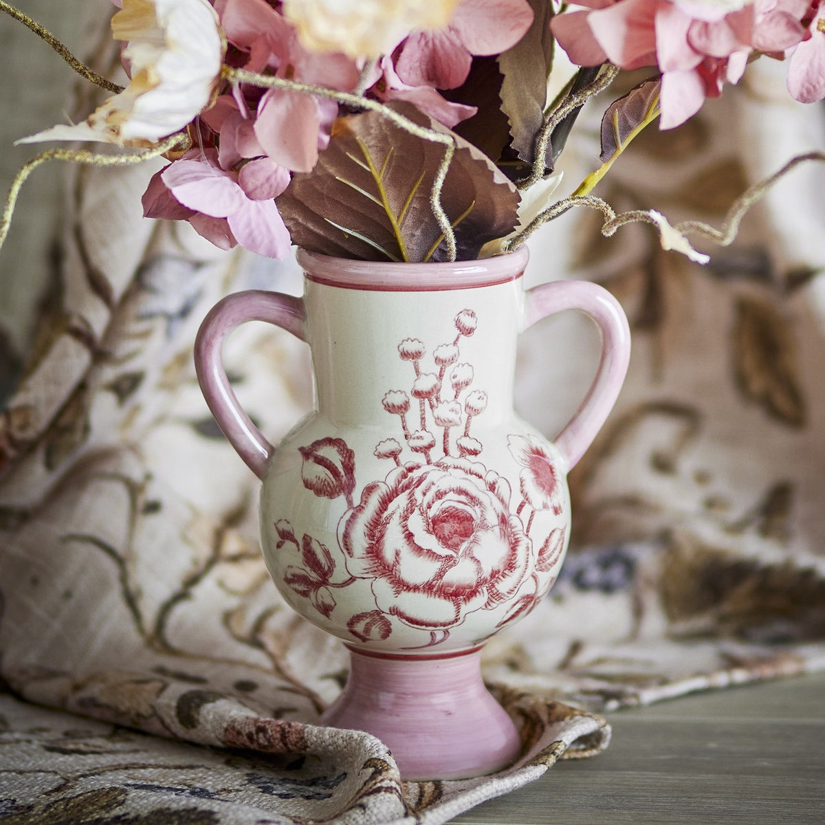 Alea Floral Stoneware Vase 21cm in pink, with round belly, rolled lip, two loop handles and pedestal foot, hand-painted rose design, shown holding pink florals against a patterned fabric backdrop.